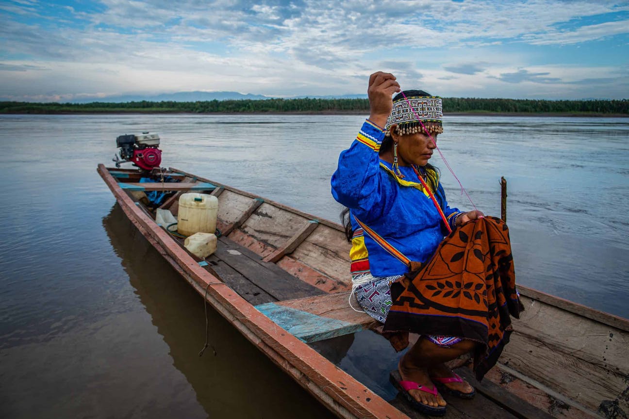 A person in traditional clothing and headgear sits in a wooden boat on a river, holding an object and performing an action, with a small motor and fuel container in the background.