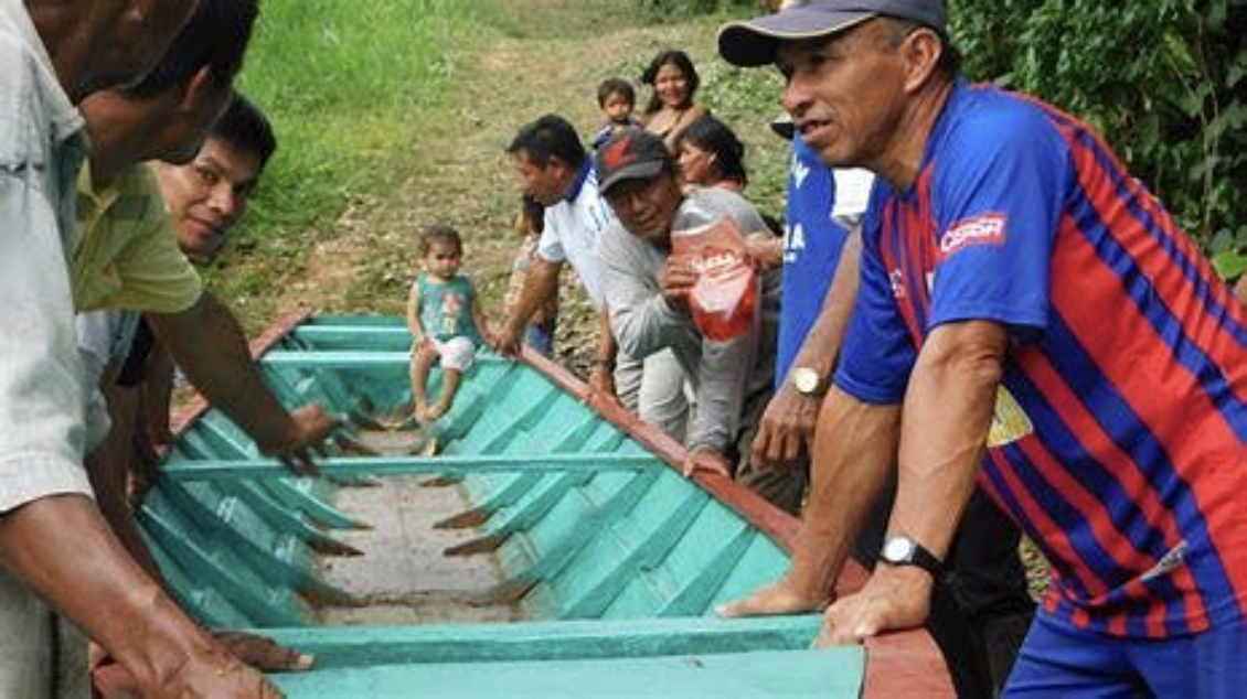 A group of people, including a man in a blue and red shirt, are standing around a wooden boat positioned on the ground, with a toddler sitting inside it.