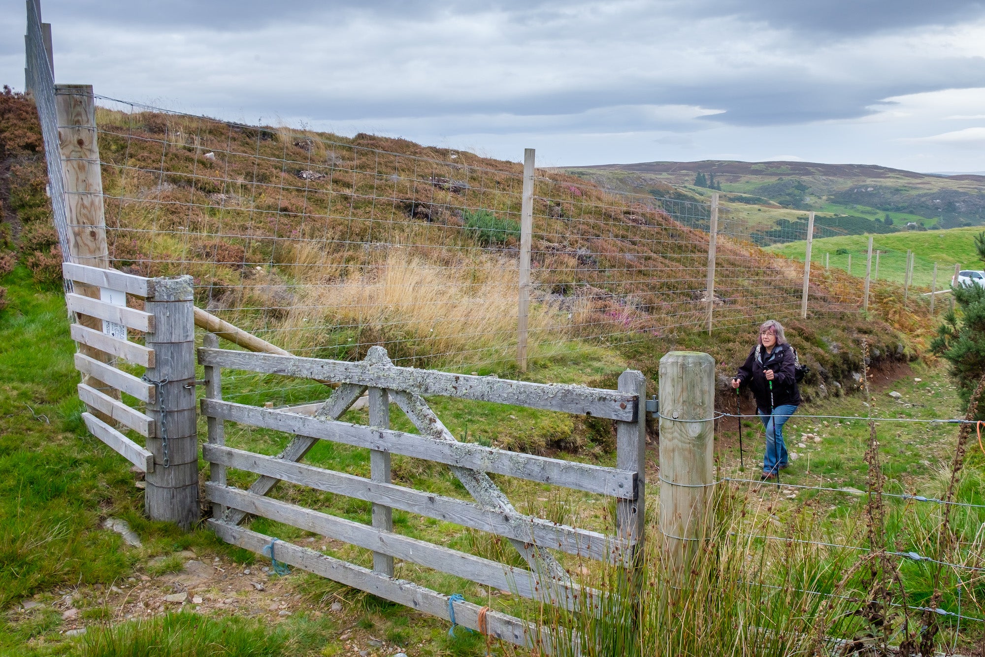 A person in outdoor clothing is seen closing a wooden gate on a rural hillside, ensuring public access to green space. The area is surrounded by grassy terrain, tall wooden fence posts, and wire fencing.