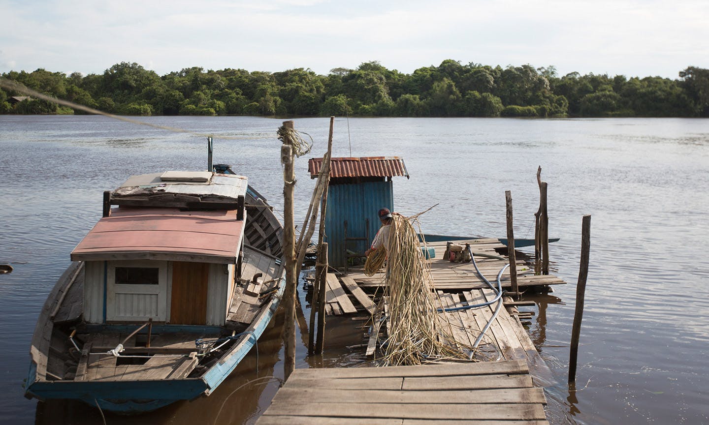 A small wooden boat with a red roofed cabin is moored at a rustic wooden dock on a calm river. Next to the boat, a person works with a pile of reeds on the dock. In the background, dense green forest lines the opposite riverbank.