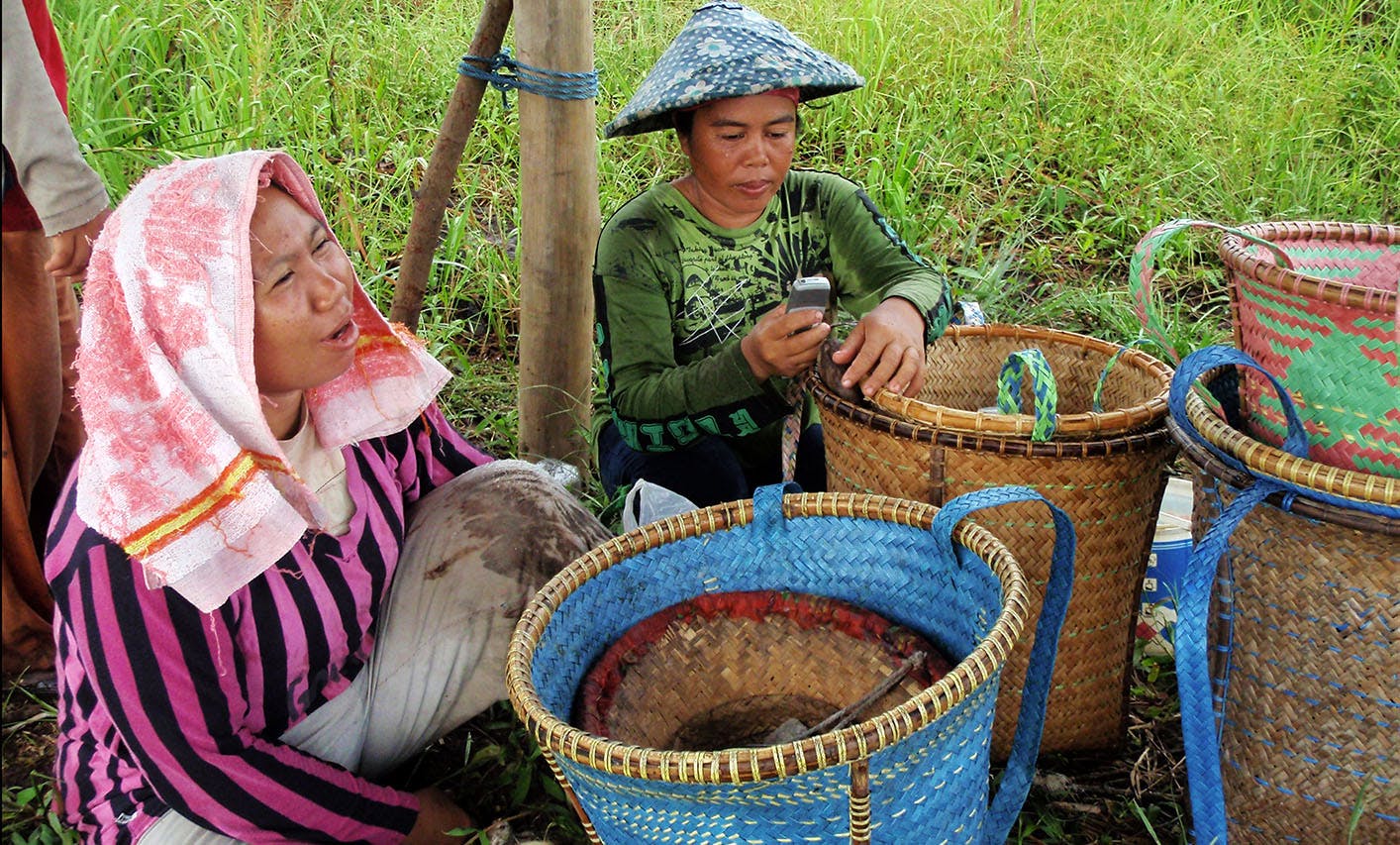 Two women sit on the ground in a grassy area. One wears a striped shirt with a towel over her head, the other a green shirt and a conical hat, using a mobile phone. They are surrounded by wicker baskets of various colours.