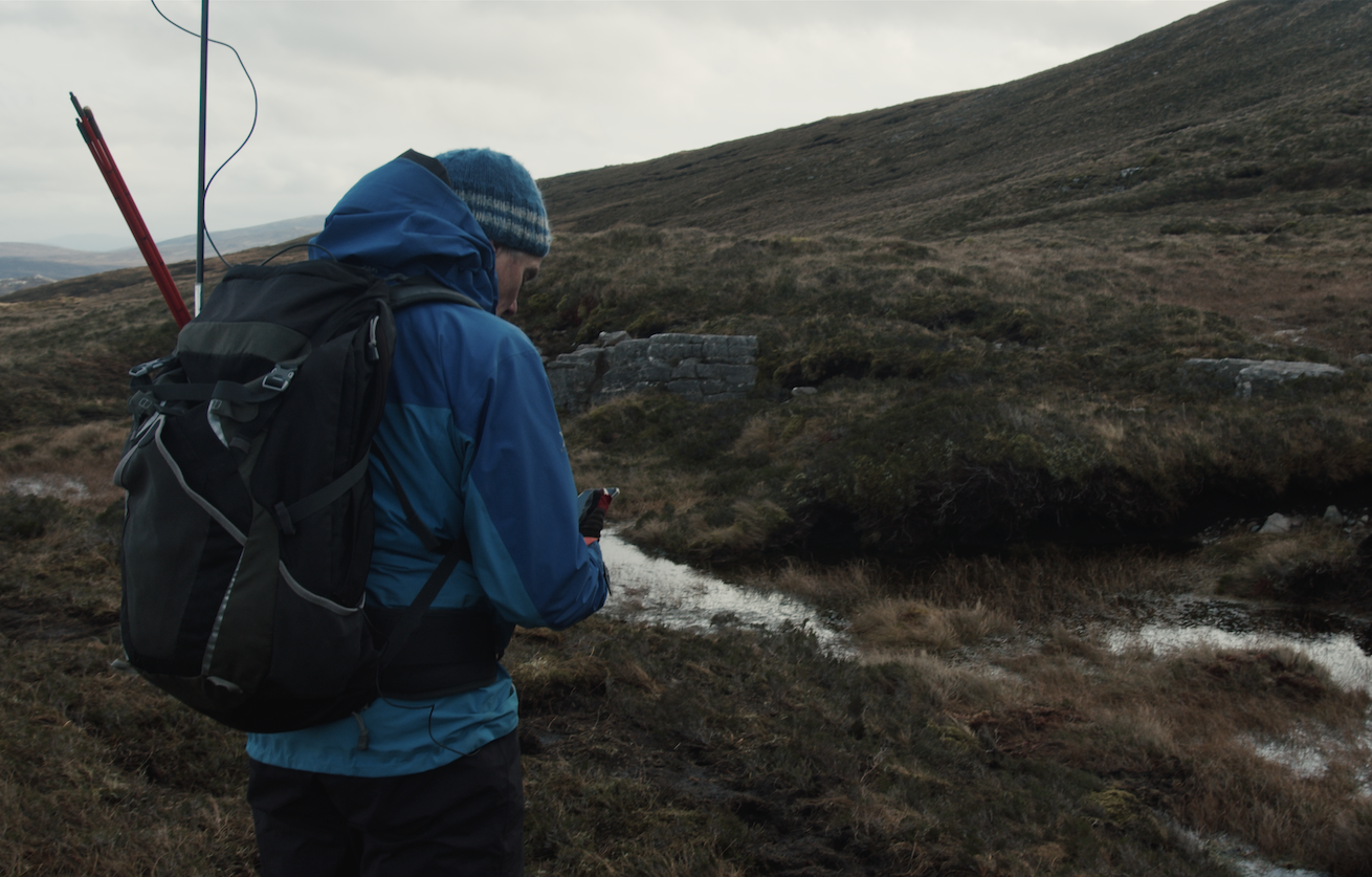 A person in a blue jacket and backpack stands on a rugged hilltop, verifying peatland restoration on their device, with cloudy skies overhead and a small stream nearby.