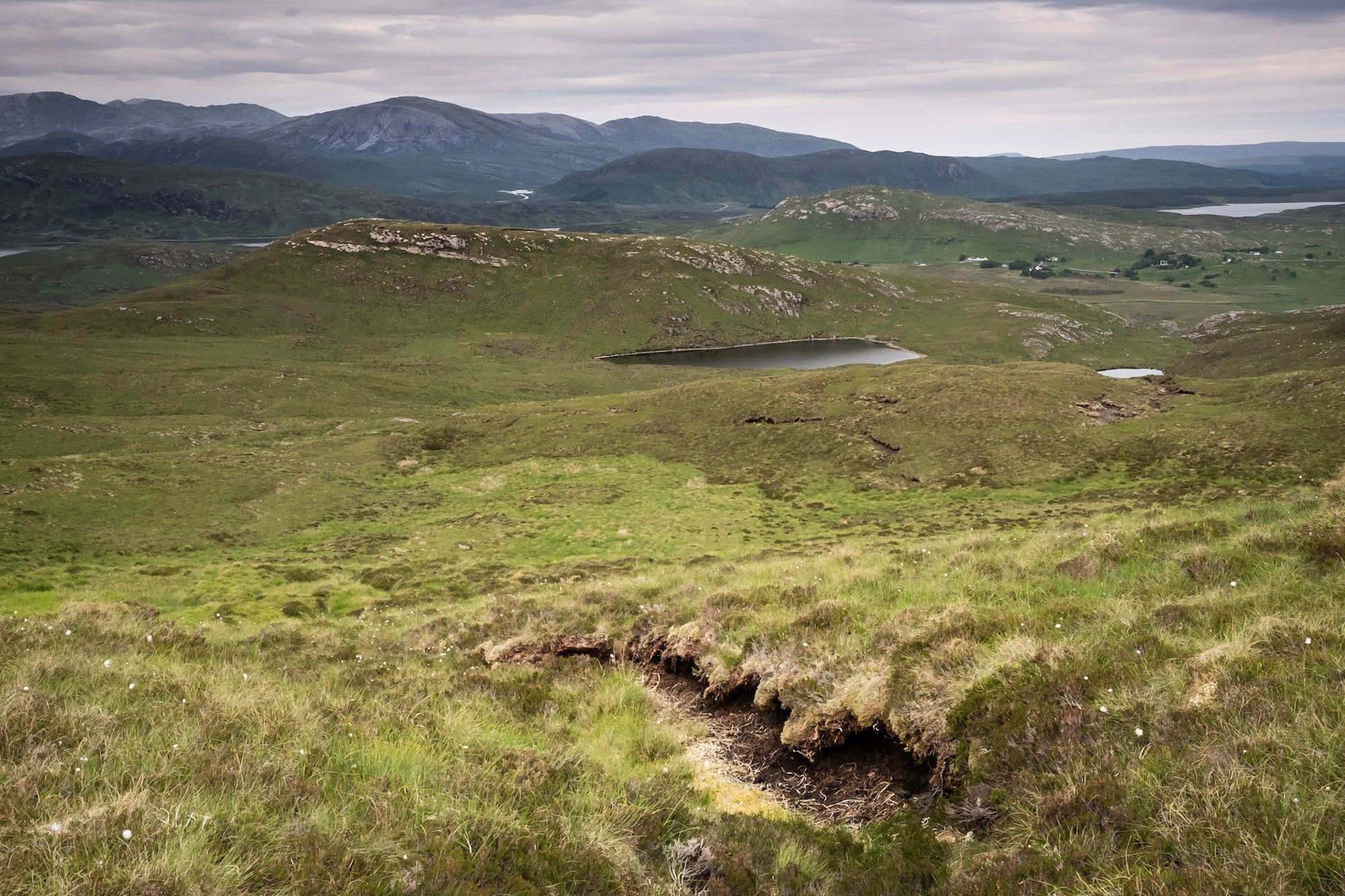 A landscape with rolling green hills, small lakes, and distant mountains under a cloudy sky; the foreground reveals exposed soil that hints at the richness of UK peatland.