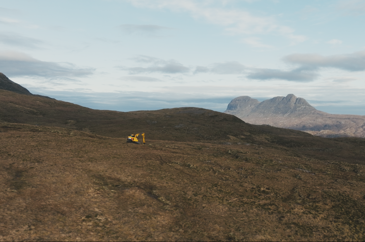 A construction vehicle works on a wide, grassy plain, contributing to peatland restoration efforts with mountains in the background under a partly cloudy sky.