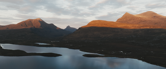 An aerial view of a mountain range at sunset reveals hills and peaks casting long shadows over a series of tranquil lakes, nestled alongside vast stretches of serene peatland.